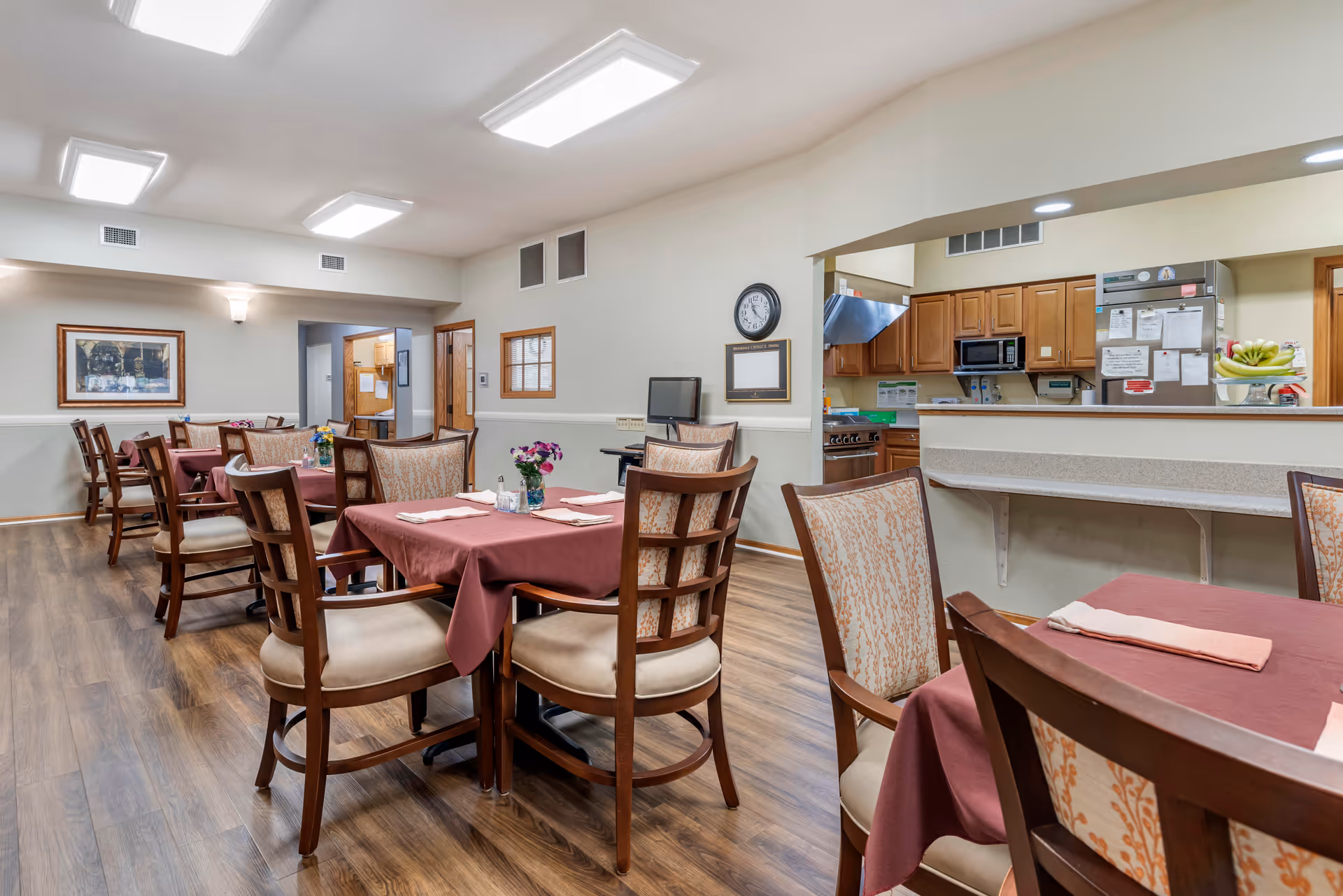 A dining room in a senior living facility with several tables covered in maroon tablecloths, each set with napkins and small flower arrangements. Wooden chairs with patterned upholstery surround the tables. The room has wood flooring, neutral-colored walls, and ceiling lights. A kitchen area with wooden cabinets, a refrigerator, and a stove is visible through a pass-through window.