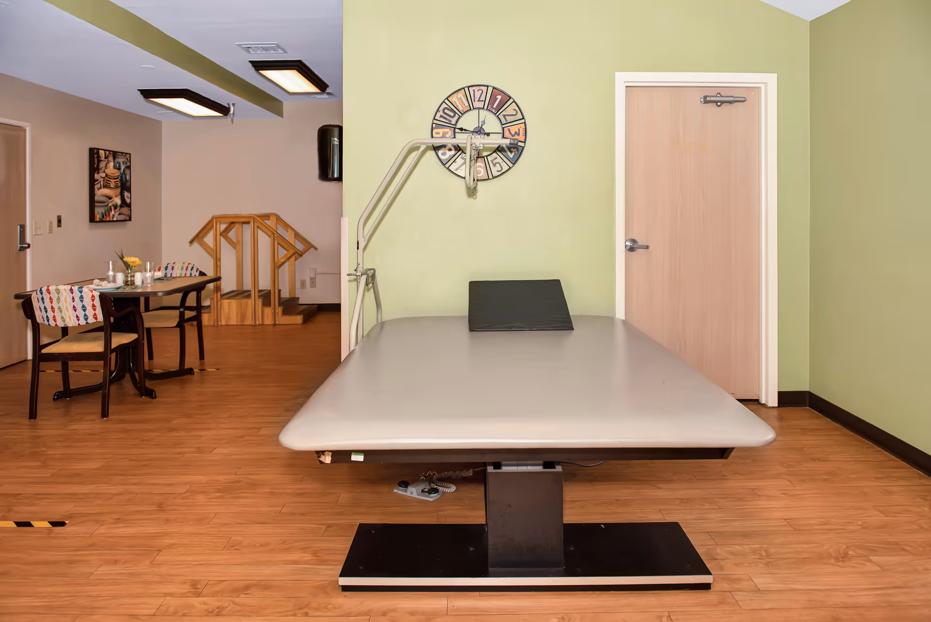 Interior room with a large medical treatment table in the foreground, a wall clock, a door, and a small dining area with chairs and a wooden step ramp in the background.