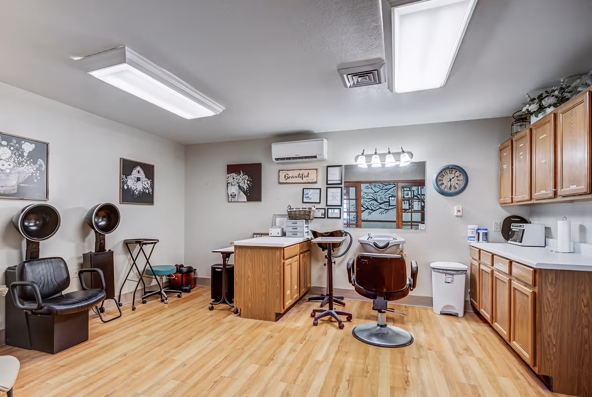 Interior view of a salon area in a senior living facility with two vintage hair dryers on the left, a central island with a chair and hair washing station, wooden cabinets on the right, and wall decorations including framed pictures and a clock.