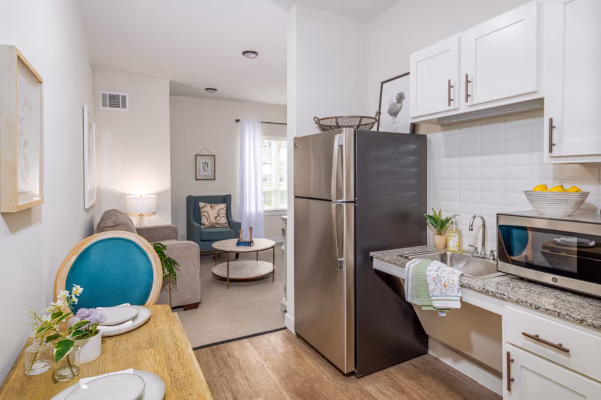Interior view of a senior living facility apartment showing a compact kitchen area with a stainless steel refrigerator, microwave, sink, and white cabinets. Adjacent to the kitchen is a small dining table with two place settings and a teal cushioned chair. In the background, a living room area is visible with a gray sofa, a teal armchair, a round coffee table, and a window with white curtains letting in natural light.