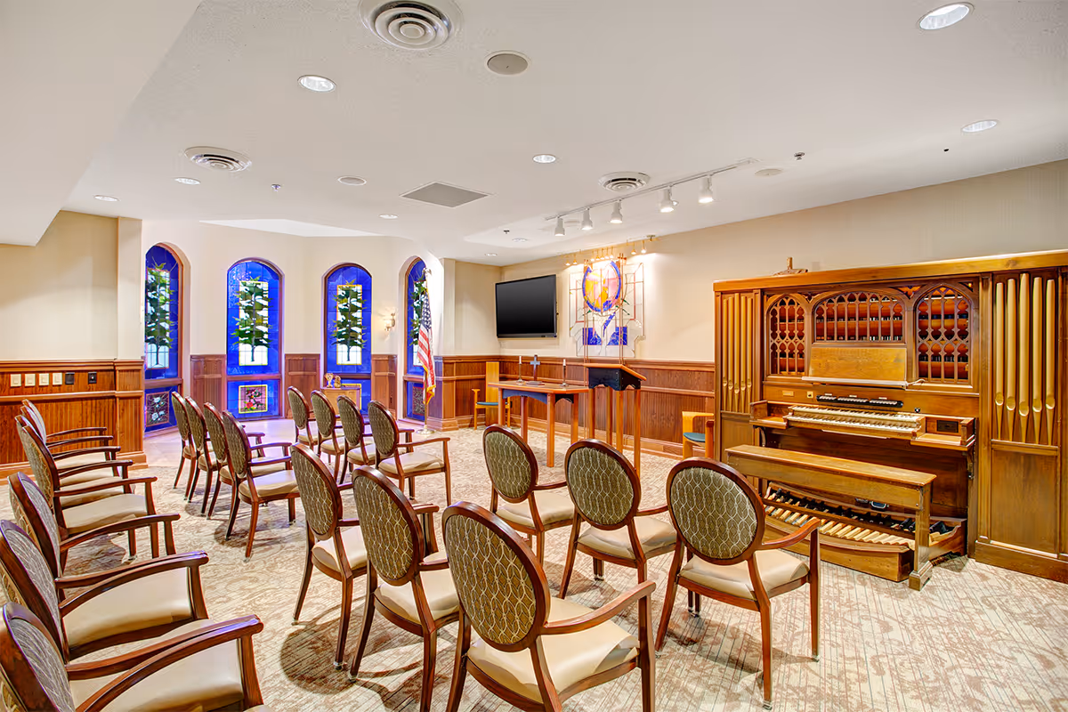 Interior view of a chapel or small worship room with rows of wooden chairs with cushioned seats facing a wooden altar and a large pipe organ. The room has stained glass windows, an American flag, a wall-mounted TV, and soft lighting from ceiling fixtures.