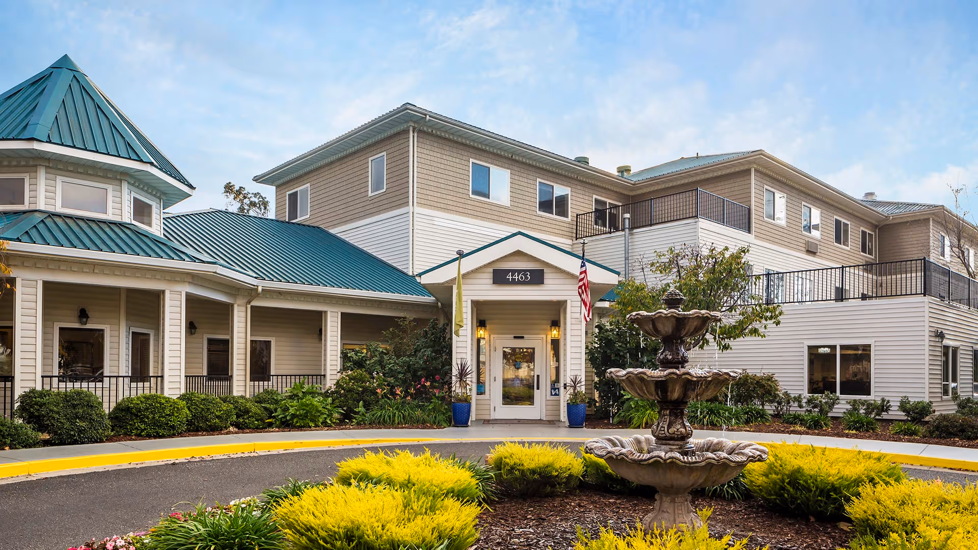 Front exterior view of a senior living facility named Atria Evergreen Valley with a circular driveway, a three-tiered fountain in the center, green shrubs, and a building with beige siding and teal metal roofs under a partly cloudy sky.