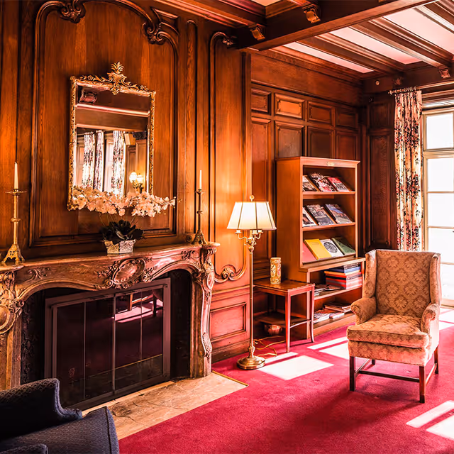 A cozy living room with rich wooden paneling and an ornate fireplace. Above the fireplace is a decorative mirror and a vase with flowers. There are two tall candlesticks on either side of the fireplace. A floor lamp stands next to a wooden bookshelf filled with magazines and books. An upholstered armchair is positioned near a window with floral curtains, and sunlight streams into the room, casting shadows on the red carpet.