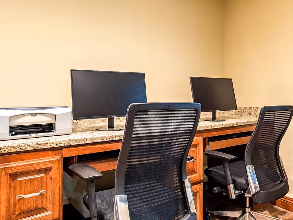 A workspace area with two black mesh office chairs in front of a wooden desk with a granite countertop. On the desk are two computer monitors and a white printer against a beige wall.