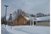 Snow-covered single-story building with a covered entrance, bare trees, and a lamp post in front.