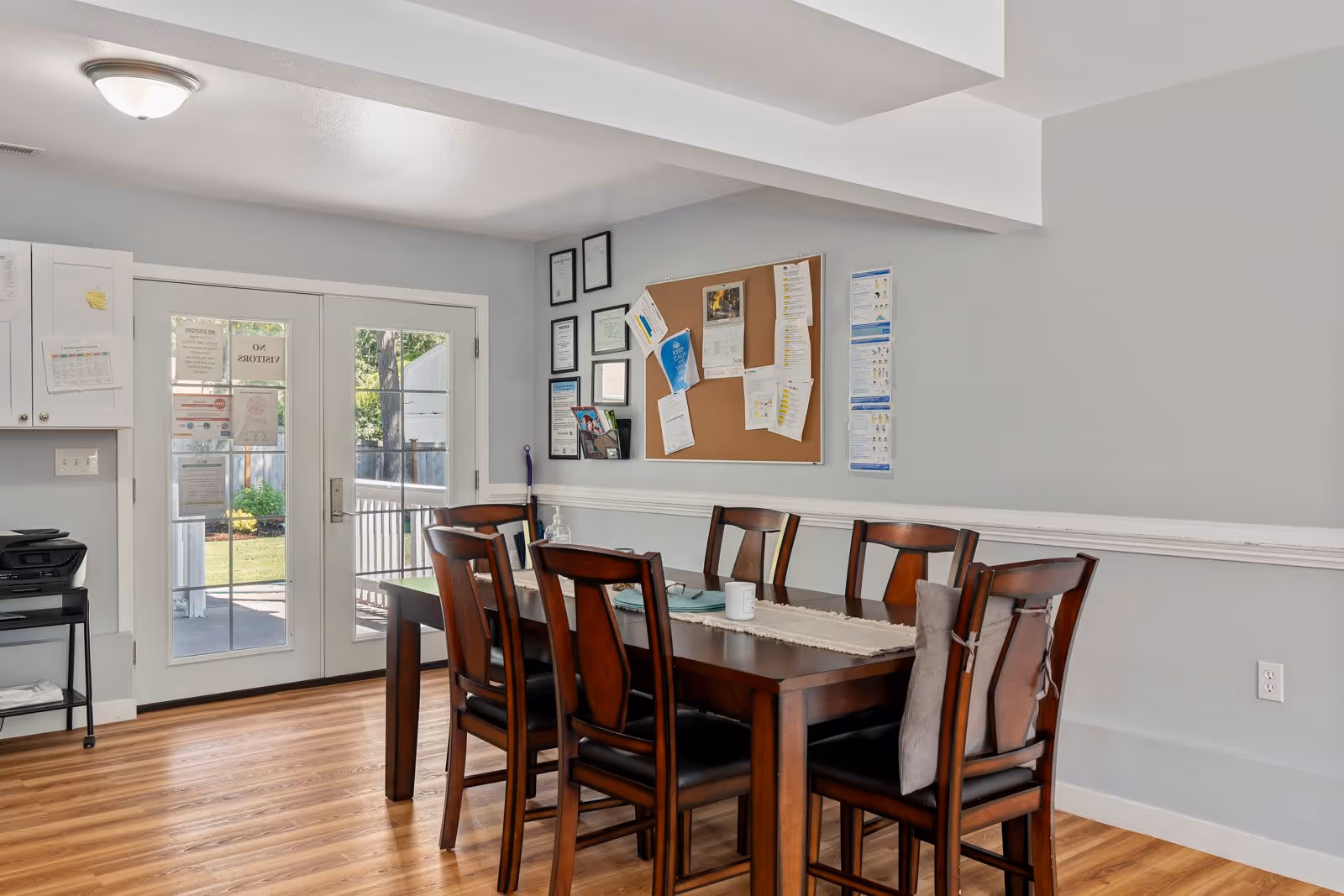 Dining area with a wooden table and six chairs, a bulletin board on the wall, and glass double doors leading outside.