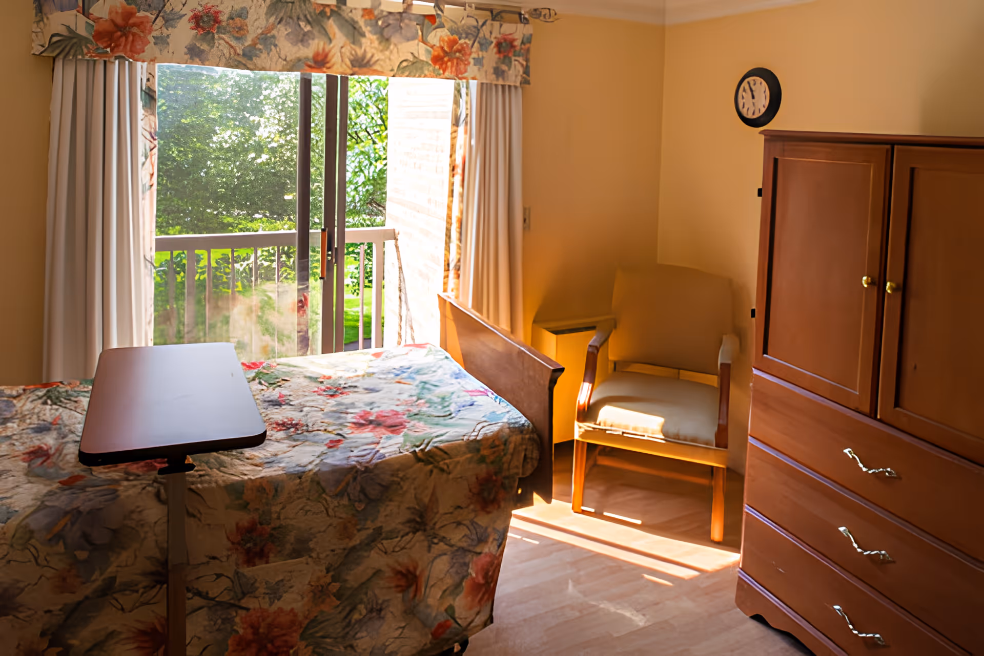 A bright retirement community bedroom with a floral bedspread on the bed, a wooden dresser, a beige armchair, and a sliding glass door leading to a balcony with green trees outside. The room has light-colored walls and a clock on the wall.
