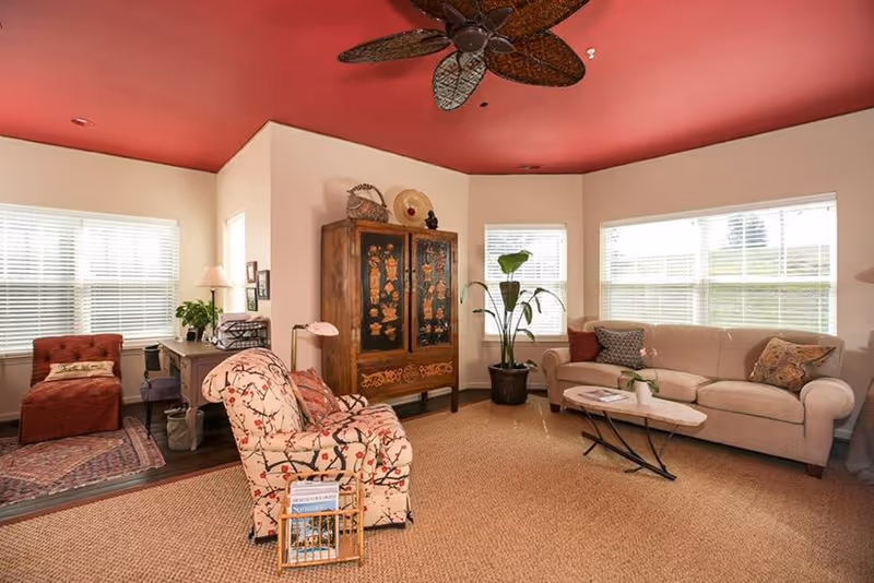 A cozy living room with a beige sofa, a patterned armchair, a wooden coffee table, a large decorative cabinet, and a ceiling fan with leaf-shaped blades. The room has large windows with white blinds, a red ceiling, and a beige carpeted floor. There is also a small desk with a lamp and a red chaise lounge in the corner.