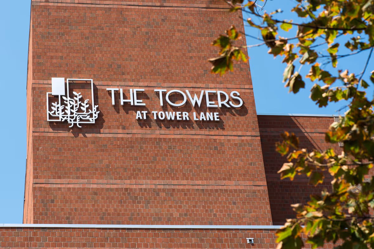 Exterior view of a brick building with a sign that reads 'The Towers at Tower Lane' against a clear blue sky, with some tree branches with green and brown leaves in the foreground.
