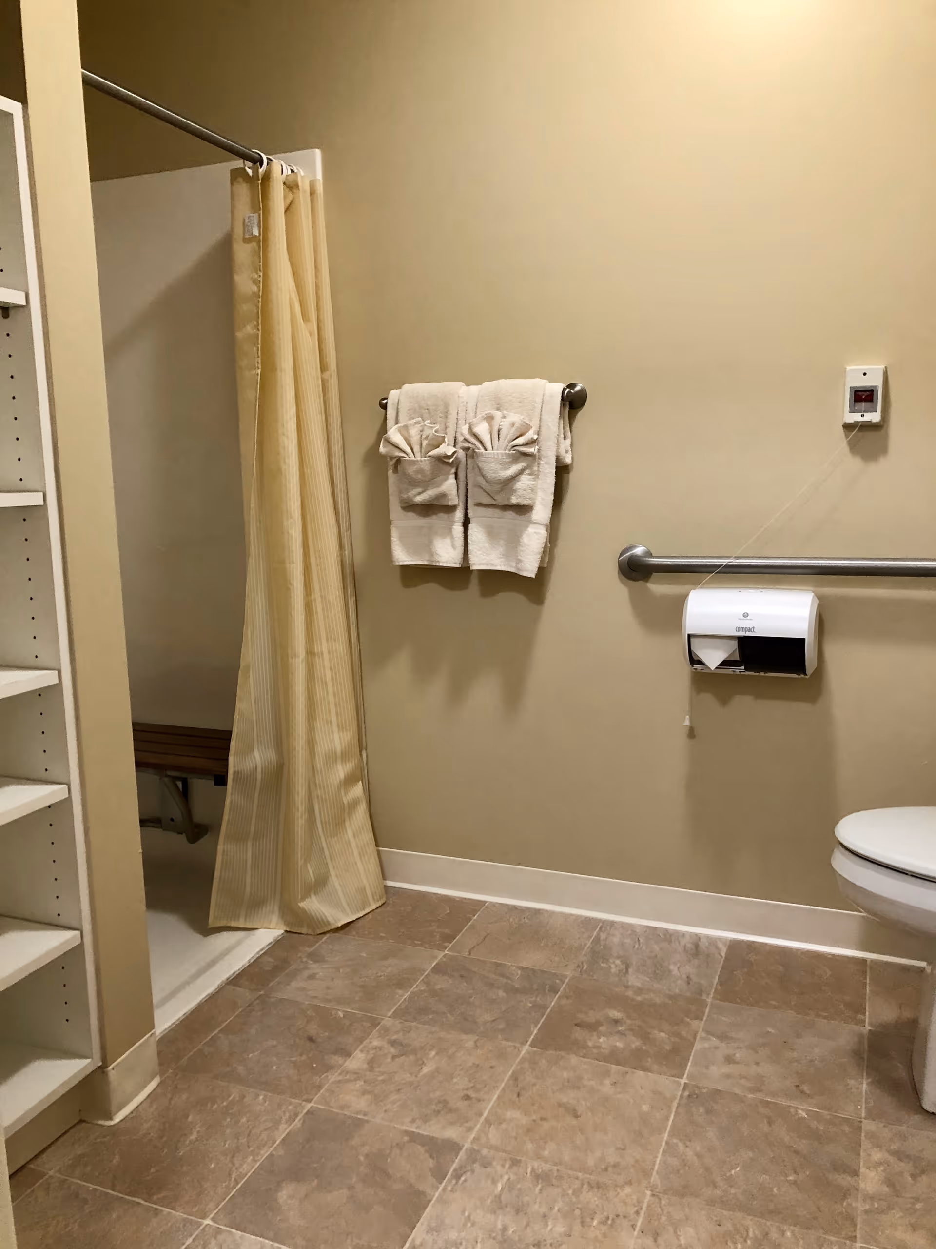 A bathroom with beige tiled floor and light beige walls. There is a shower area with a beige striped curtain on the left, a towel rack with two neatly folded white towels, a grab bar, a toilet paper dispenser, and a portion of a toilet visible on the right side.