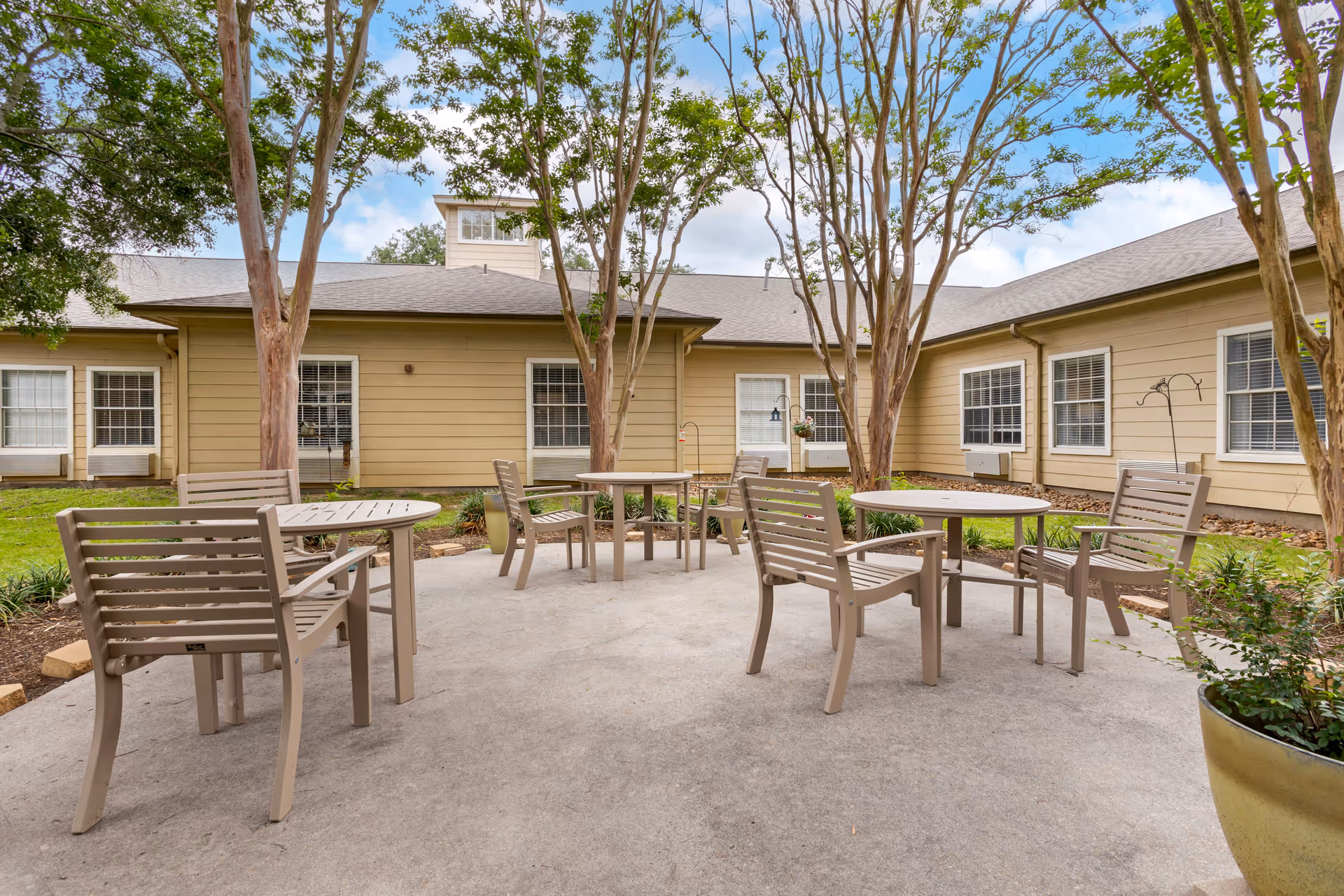Outdoor patio area at Windermere Estates Senior Living with several beige chairs and round tables arranged on a concrete surface, surrounded by trees and beige building walls with windows.