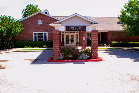 Front exterior view of a single-story brick building with a covered entrance supported by brick columns. The sign above the entrance reads 'Emeritus Senior Living'. There are trees and shrubs around the building and a parking area in front.