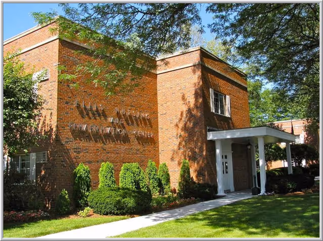 Brick two-story building front with a covered entrance and manicured shrubs under trees.