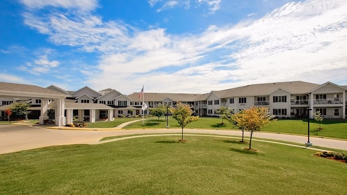 Exterior view of Curtis Creek Senior Living facility showing a large, two-story building with multiple windows and balconies, surrounded by a well-maintained lawn with small trees and a driveway. The sky is partly cloudy with blue patches.