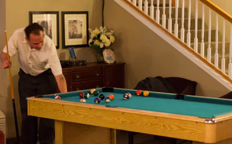 A man playing pool in a room with a wooden pool table. Behind him is a wooden cabinet with a vase of flowers and framed pictures on the wall. A staircase with white railings is visible in the background.