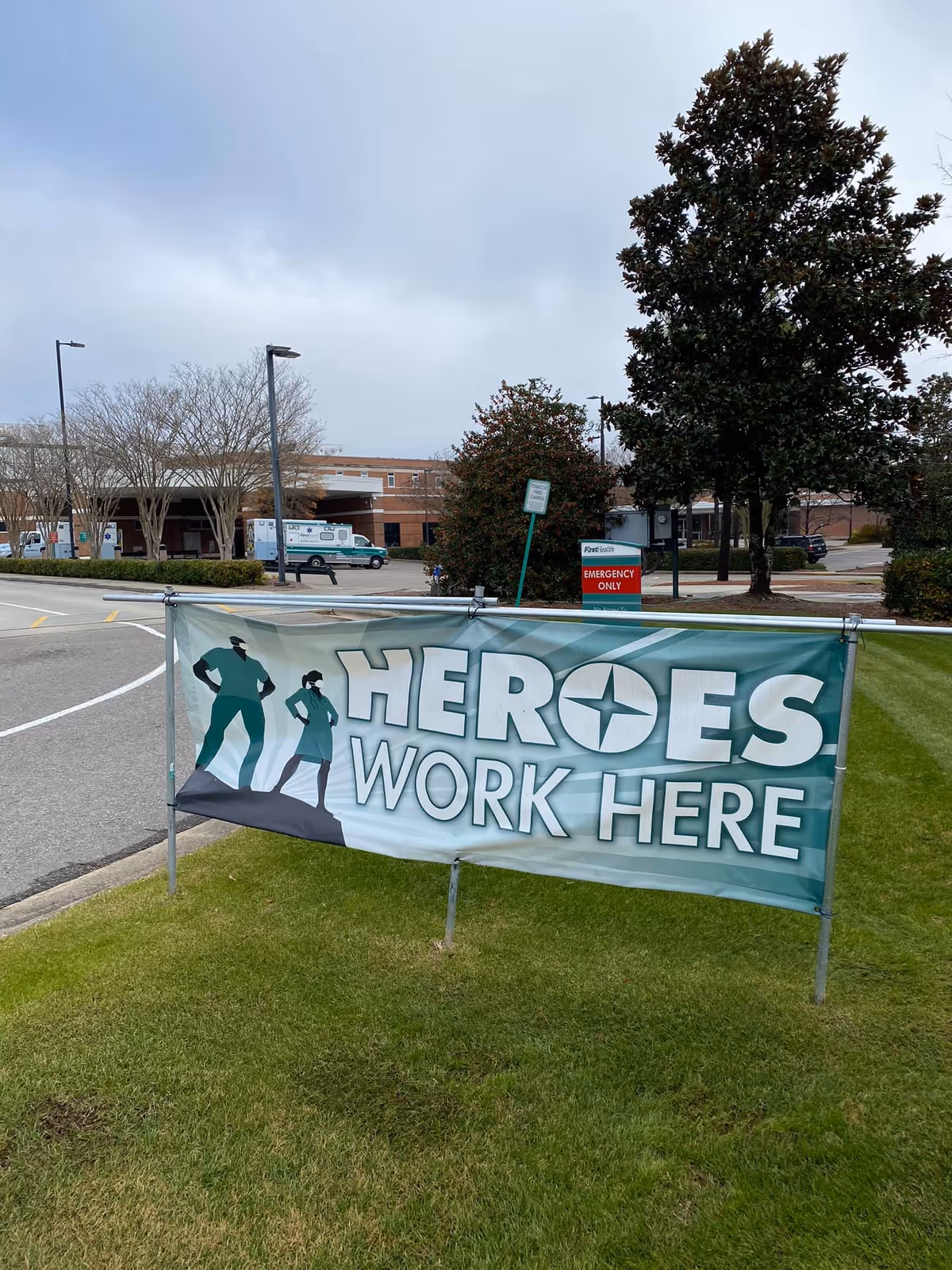A lawn banner reading "HEROES WORK HERE" in front of a rehabilitation and living center entrance with ambulances and the building visible behind it.