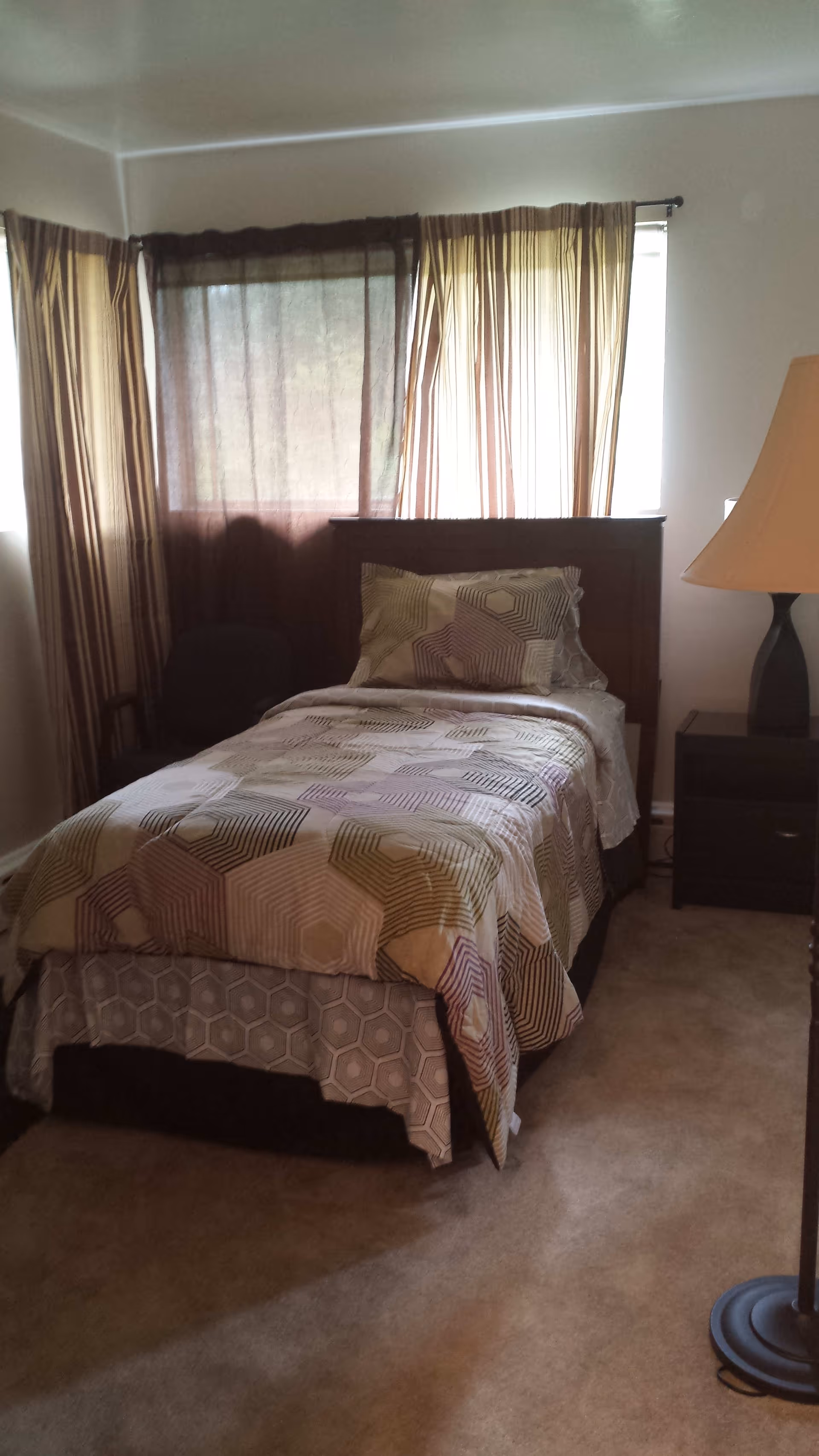 Single-occupancy bedroom with a twin bed under curtained windows, patterned bedding, a nightstand and lamp.