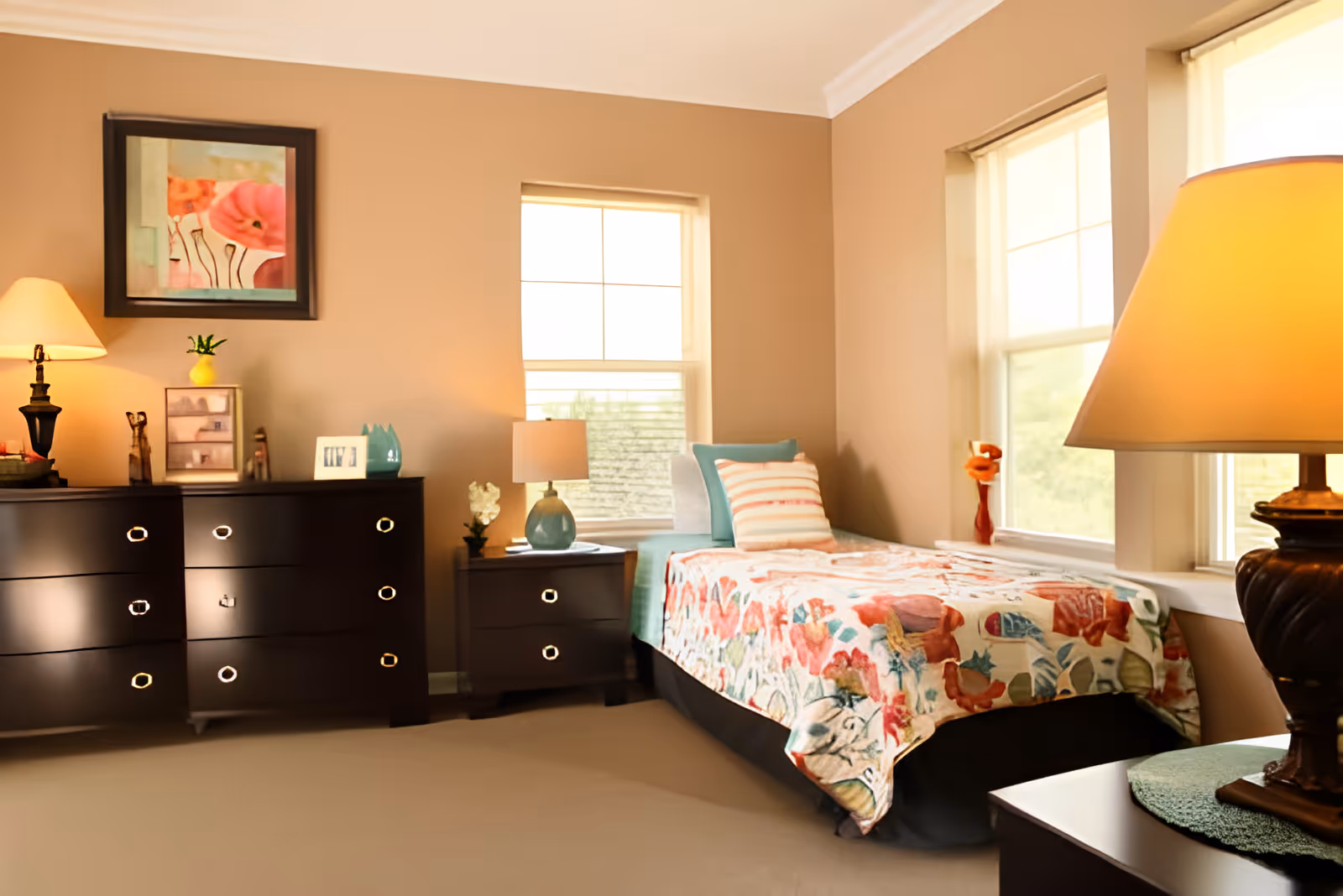 A cozy bedroom with beige walls, a single bed covered with a colorful floral bedspread, and two pillows. The room has two windows letting in natural light. There are two dark wood dressers, one with a lamp, framed photo, and decorative items, and the other with a lamp and small plant. A framed floral painting hangs on the wall above one dresser.