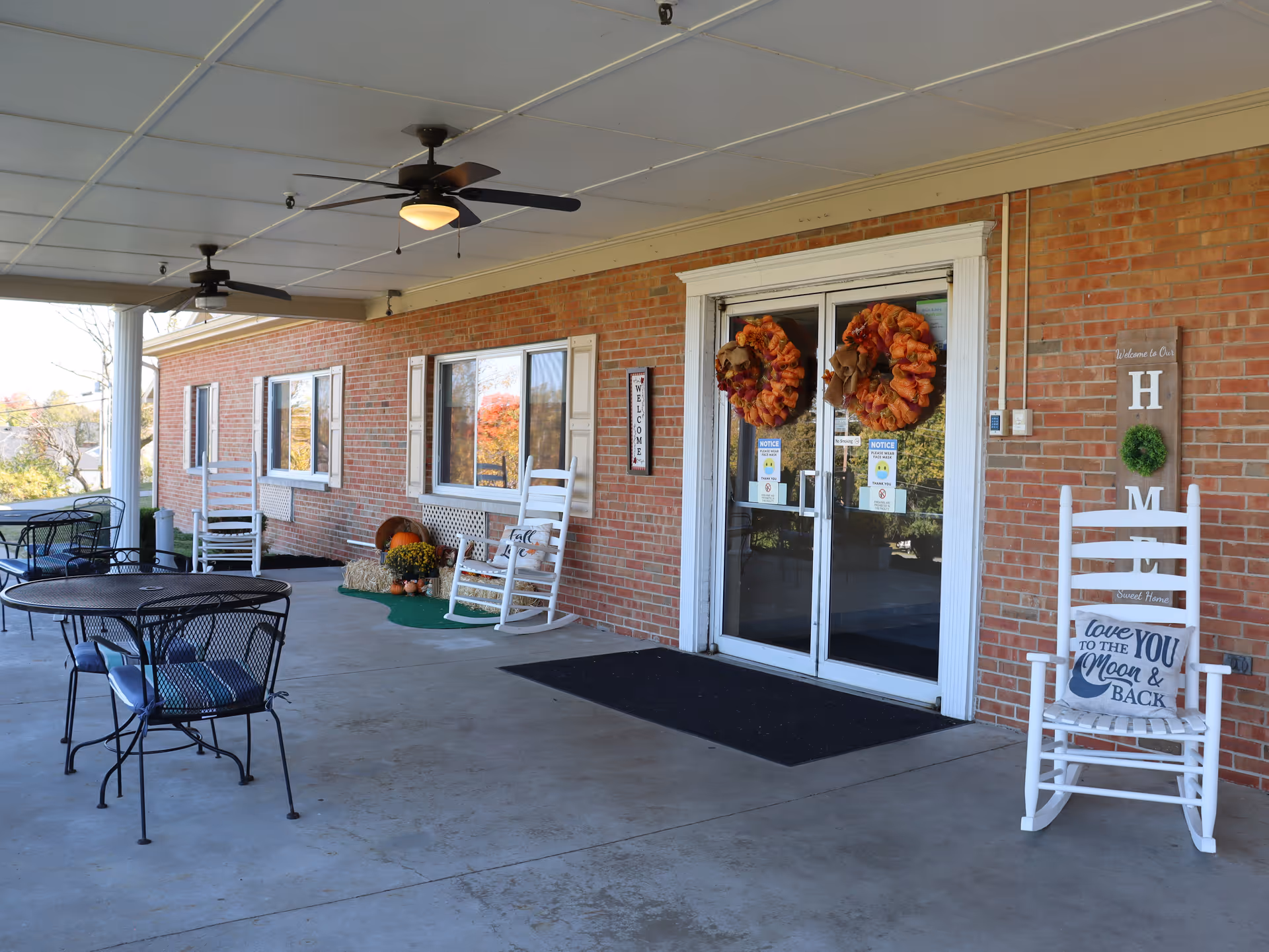 Covered outdoor patio area of a brick building with white rocking chairs, black metal tables and chairs, ceiling fans, and fall-themed wreaths on glass double doors. There are decorative signs and plants near the entrance.