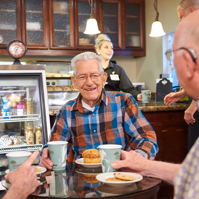 An elderly man wearing glasses and a plaid shirt sits at a round table with a muffin and a cup in front of him, smiling. Two other elderly people are partially visible at the table, holding cups and plates with cookies. In the background, a young woman in a black uniform stands behind a counter with baked goods and beverages, smiling. The setting appears to be a cozy cafe or dining area with wooden cabinets and hanging pendant lights.