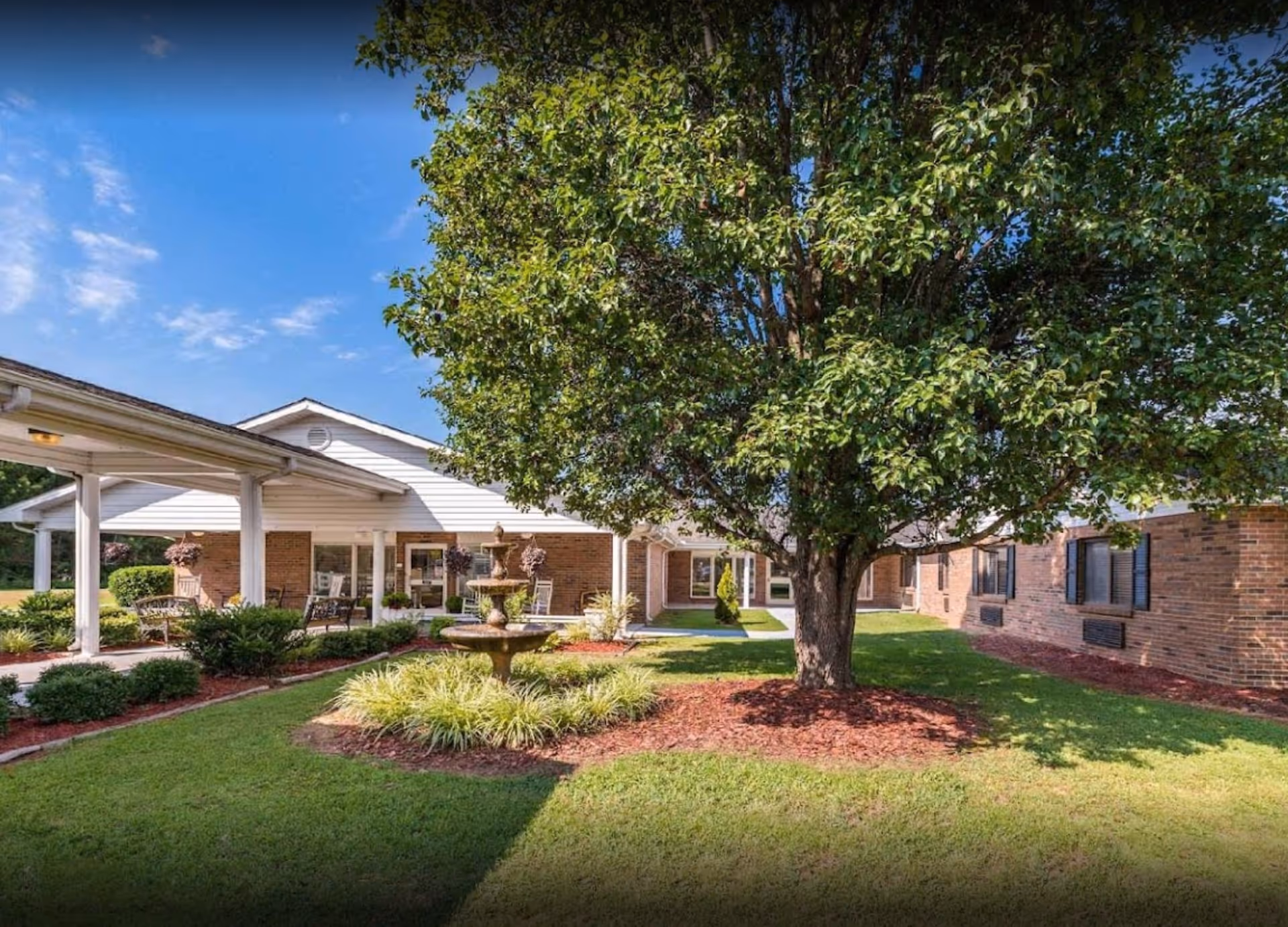 Exterior view of a senior living facility with a fountain, tree, and covered walkway.