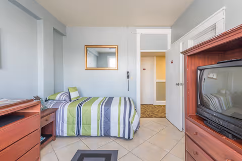 Small furnished bedroom with a single striped green-and-gray bed, wooden dressers, and a CRT television.