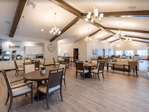Spacious communal dining and lounge area with round wooden tables and chairs, a kitchenette on the left, and a vaulted ceiling with exposed beams and chandeliers.