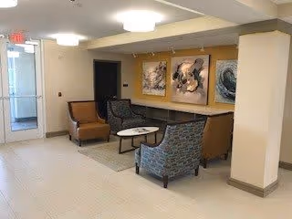 A seating area in a senior living facility with four armchairs arranged around two small round tables on a rug. The walls are decorated with three abstract paintings, and there is a glass door leading outside on the left side.