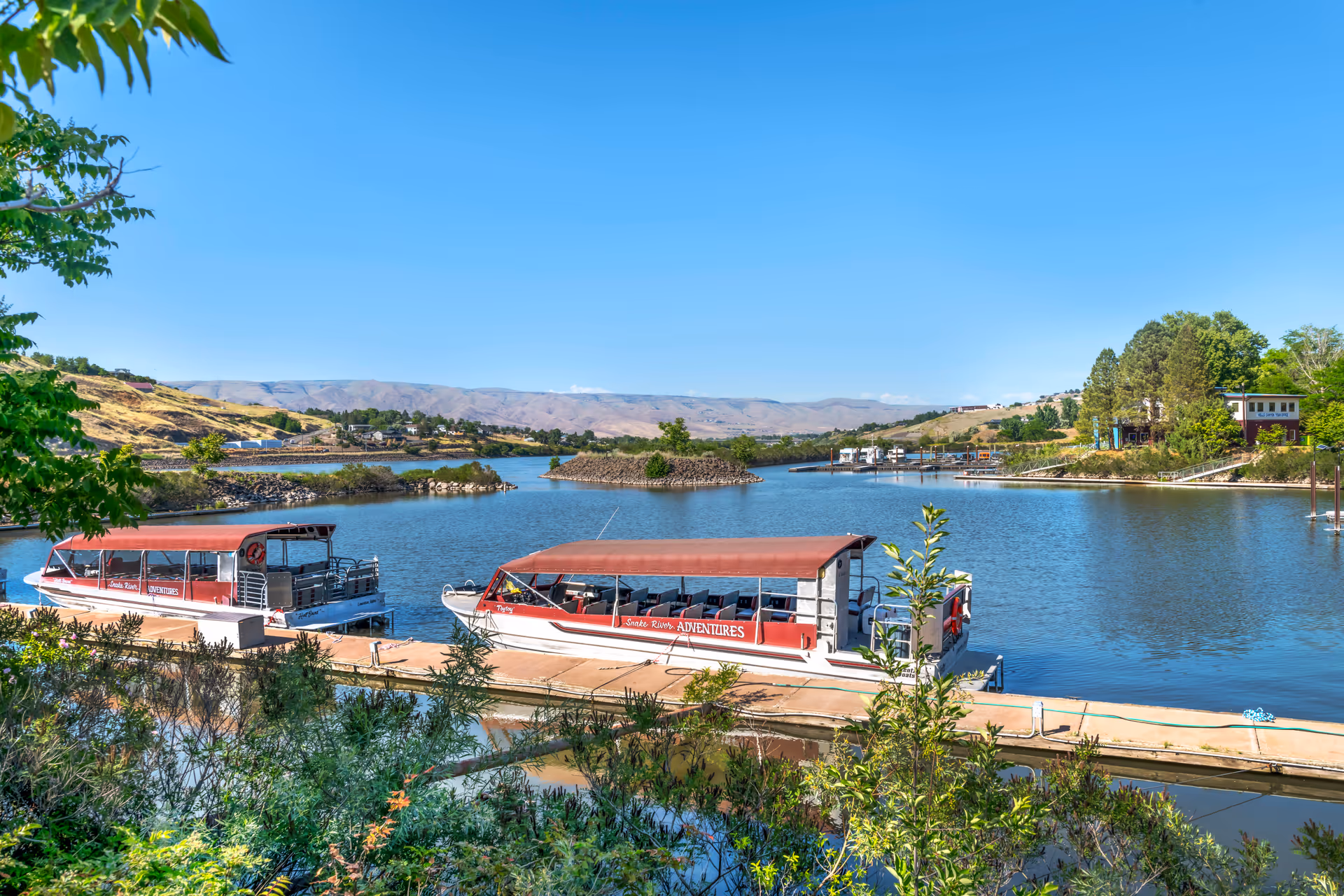 A scenic view of a calm river or lake with two red and white covered boats docked at a wooden pier. The surrounding area includes green trees and shrubs, with hills and a clear blue sky in the background.