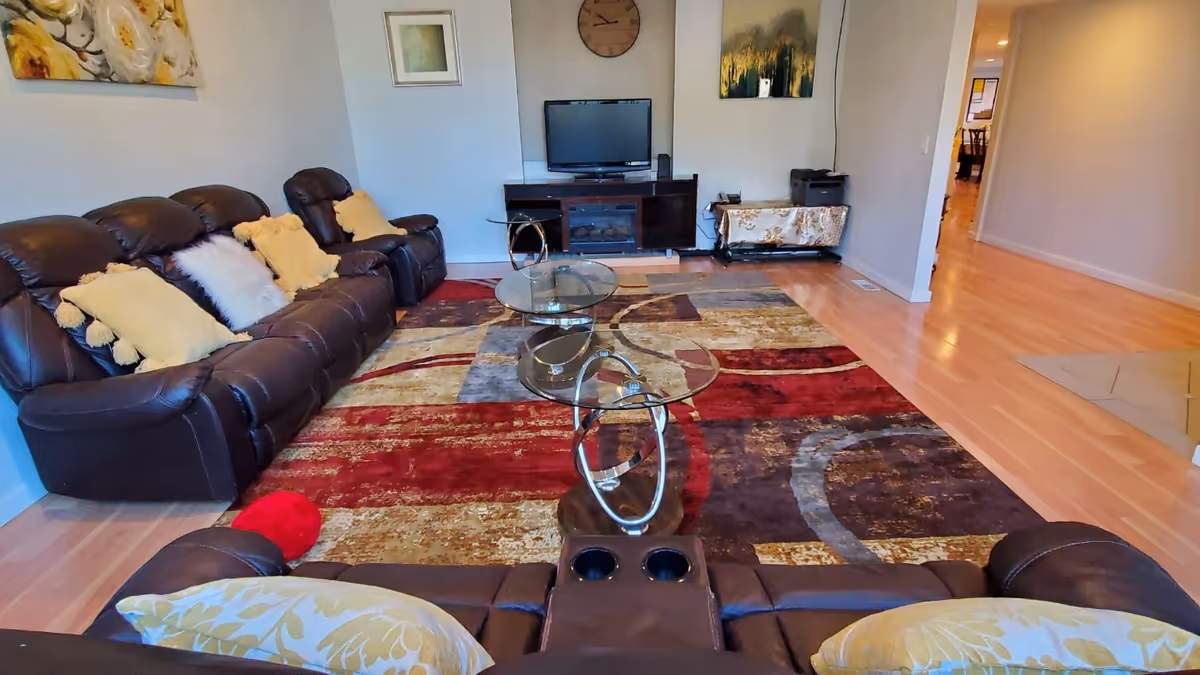 A living room with two dark brown leather sofas adorned with yellow and white pillows, a colorful area rug with red, beige, and brown patterns, two glass coffee tables with metal bases, a TV on a wooden stand, wall art, and a clock. The room has light wood flooring and a hallway visible in the background.