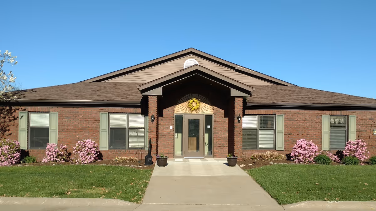 Front exterior view of a single-story brick building with a peaked roof, green window shutters, and a glass entrance door. There are flower beds with pink blooming bushes on either side of the entrance and a concrete walkway leading up to the door under a clear blue sky.