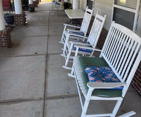 A covered outdoor patio area with a concrete floor featuring white wooden rocking chairs and a bench with colorful cushions. There are brick pillars supporting the roof and a white table in the background.