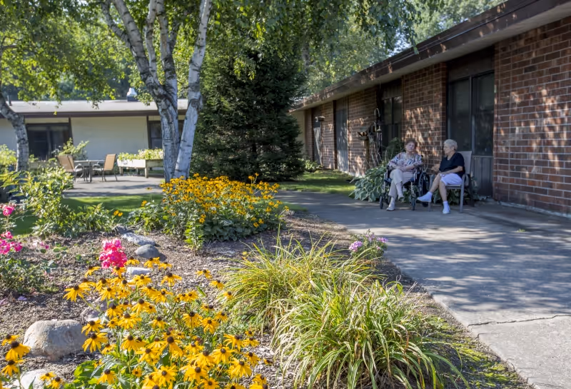 Two elderly women sit and chat on chairs outside a brick senior living building beside a flower-filled garden and walkway.