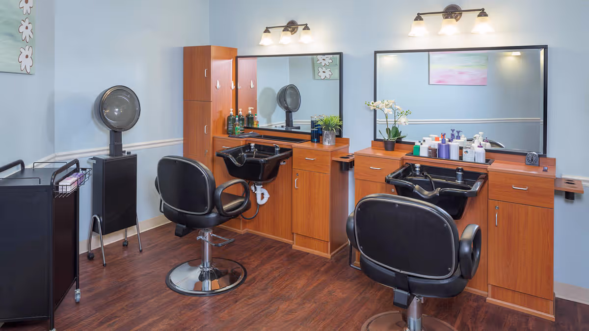Interior view of a hair salon area with two black salon chairs in front of wooden cabinets and sinks. Each station has a large mirror with lights above, various hair care products on the counters, and a hair dryer on a stand to the left. The walls are painted light blue and the floor is dark wood.