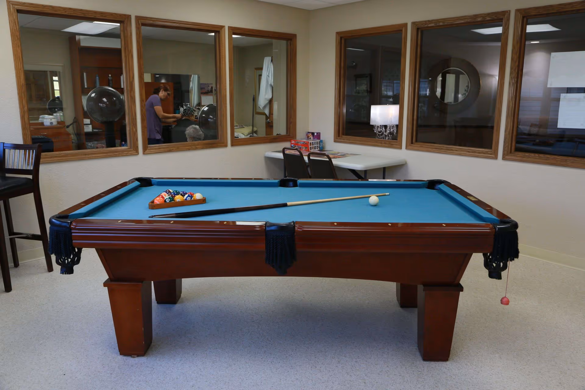 A pool table with blue felt in a recreation room with chairs, a table and windows into adjacent rooms.