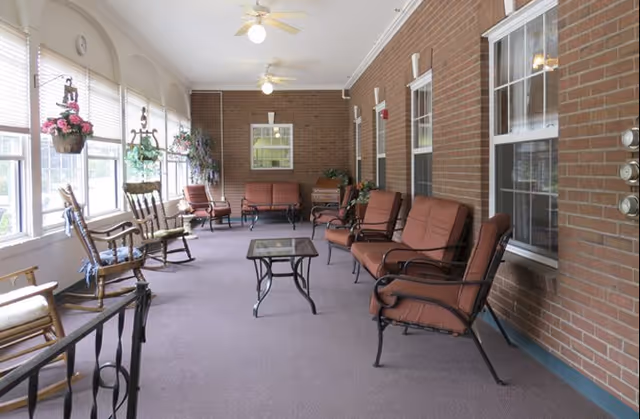 Enclosed sunroom lounge with cushioned armchairs and rocking chairs arranged around small tables along a wall of windows.