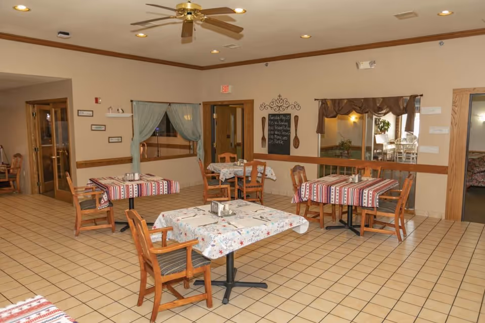A bright communal dining area with several small tables covered in patterned tablecloths and wooden chairs on a tiled floor.