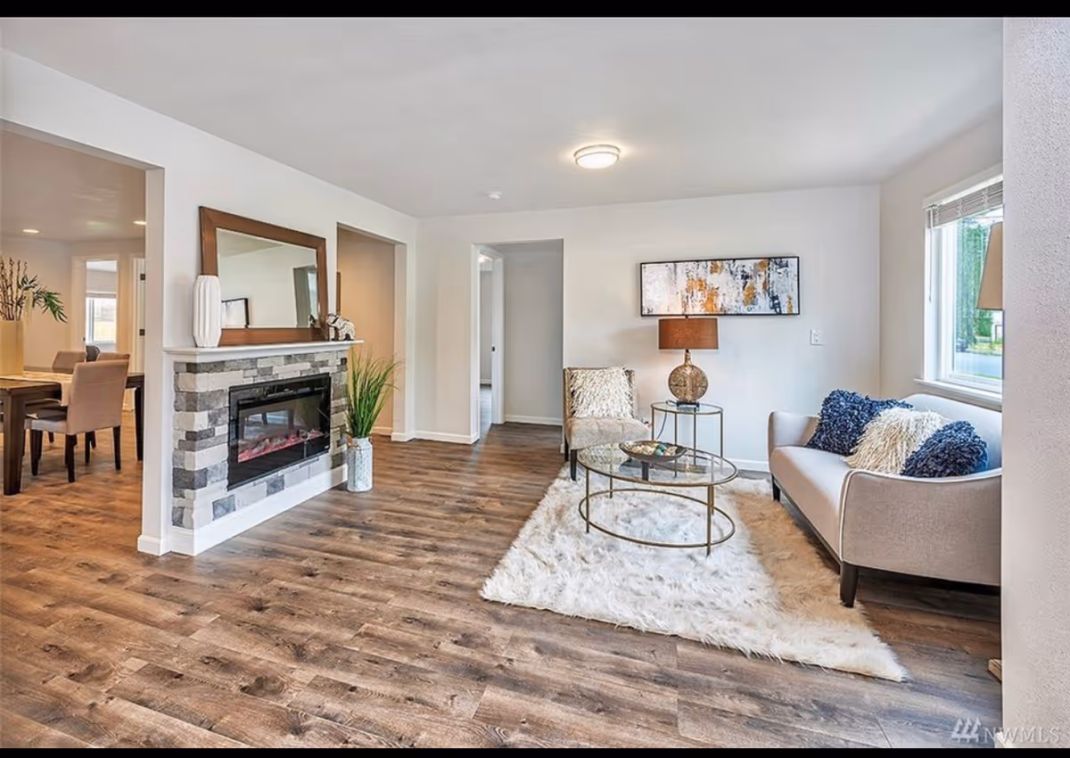 A bright and modern living room with wood flooring, a stone fireplace with a mirror and decorative items on the mantel, a light gray sofa with blue and white pillows, a beige armchair, a round glass coffee table on a white fluffy rug, a side table with a lamp, abstract wall art, and a window letting in natural light. Adjacent to the living room is a dining area with a wooden table and upholstered chairs.