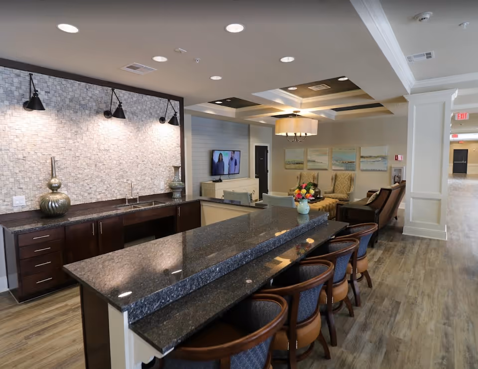 Interior view of a senior living facility common area featuring a long granite countertop with several chairs, a small sink with decorative vases on the counter, a seating area with armchairs and a coffee table with flowers, a wall-mounted TV, and soft lighting fixtures on the ceiling and wall.