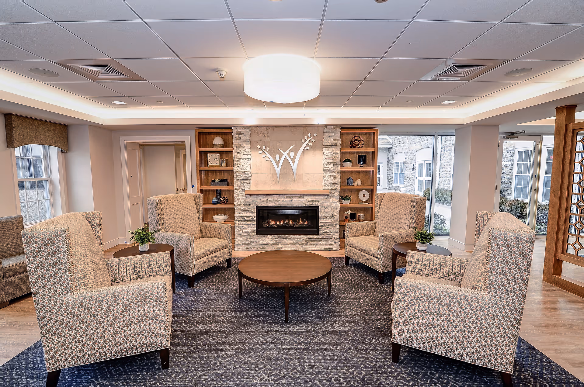 A cozy living room area with four beige patterned armchairs arranged around a round wooden coffee table on a blue patterned rug. The room features a modern stone fireplace with a decorative emblem above it, flanked by wooden shelves with various decorative items. Large windows and glass doors allow natural light to enter the space.