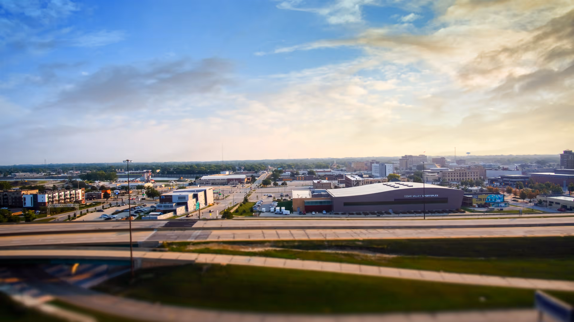 A wide aerial view of a cityscape with multiple buildings, roads, and parking lots under a partly cloudy sky during daylight.