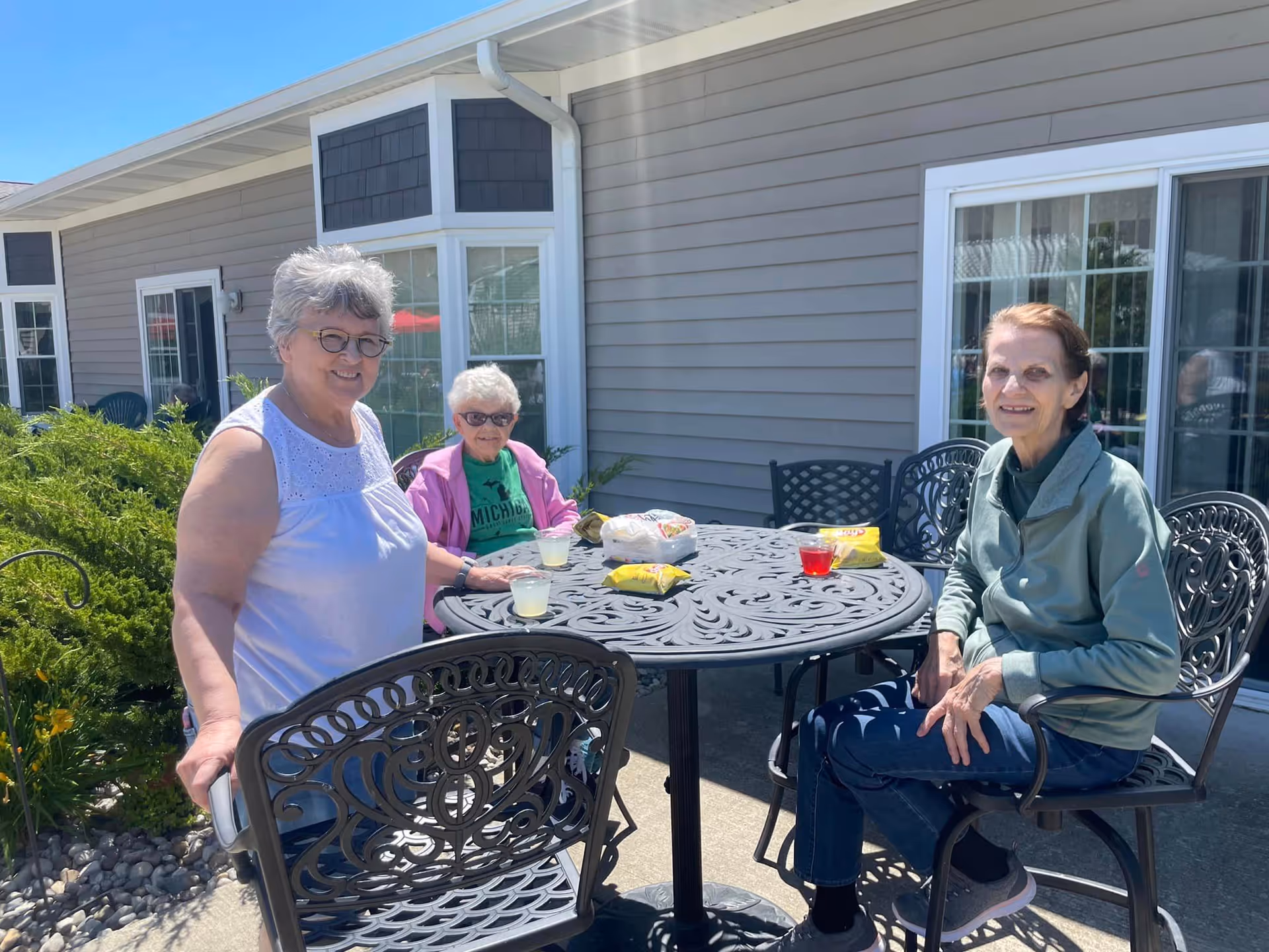 Three elderly women sitting and standing around a black metal outdoor table with snacks and drinks on it, outside a building with beige siding and large windows on a sunny day.