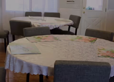 Round tables covered with lace tablecloths and place mats surrounded by upholstered chairs in a dining area with a door and curtained windows.