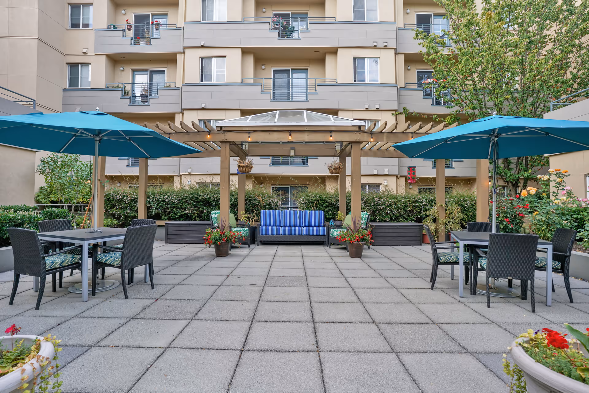 Outdoor patio area with two tables each shaded by a large blue umbrella, surrounded by black wicker chairs with patterned cushions. In the center, there is a wooden pergola with hanging plants and a blue striped cushioned bench underneath. The patio is paved with large square tiles and bordered by greenery and flowering plants. In the background, a multi-story residential building with balconies is visible.