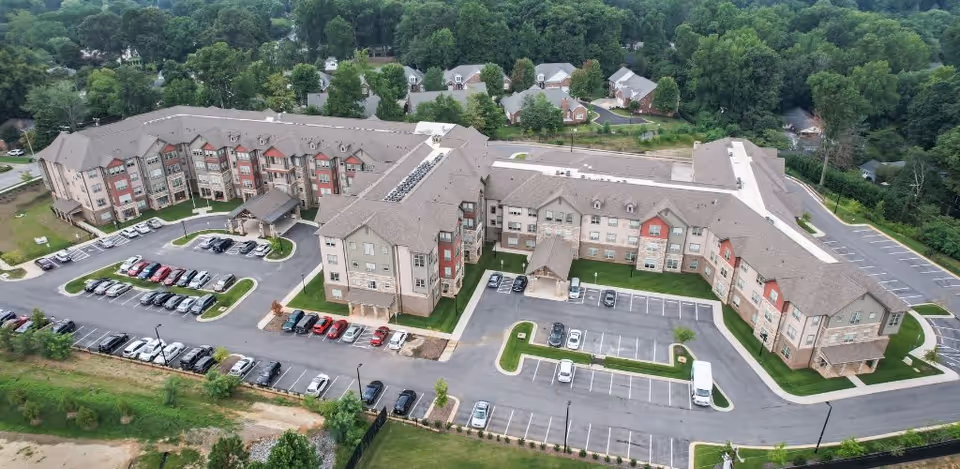 Aerial view of a large senior living facility named Harmony at Greensboro, showing a multi-wing building with a parking lot filled with cars, surrounded by trees and residential houses in the background.