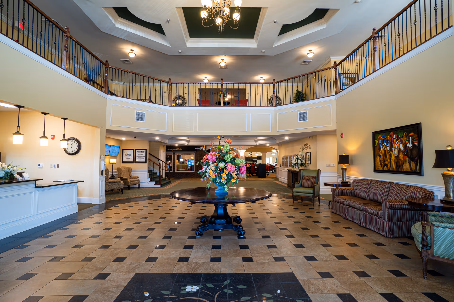 Spacious senior living facility lobby with a large round table holding a colorful floral arrangement in the center. The room features a high ceiling with a chandelier, a second-floor balcony with railing, a reception desk on the left, and seating areas with chairs and a striped sofa on the right. The floor has a patterned tile design, and there is a painting of horses on the right wall.