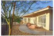 Curved paved walkway beside a beige building with large windows, desert landscaping, a tree, and decorative rocks under a clear sky.