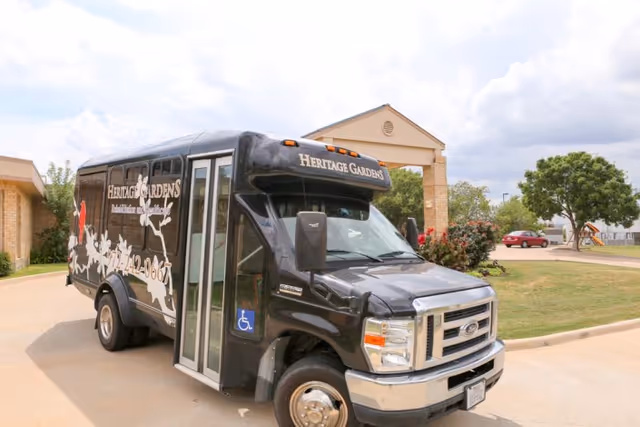 A black Heritage Gardens Rehabilitation & Healthcare shuttle bus parked on a driveway in front of a building entrance with a covered portico. The bus has white graphics and text, including a phone number and a wheelchair accessibility symbol. There are trees, bushes, and a red car visible in the background under a partly cloudy sky.