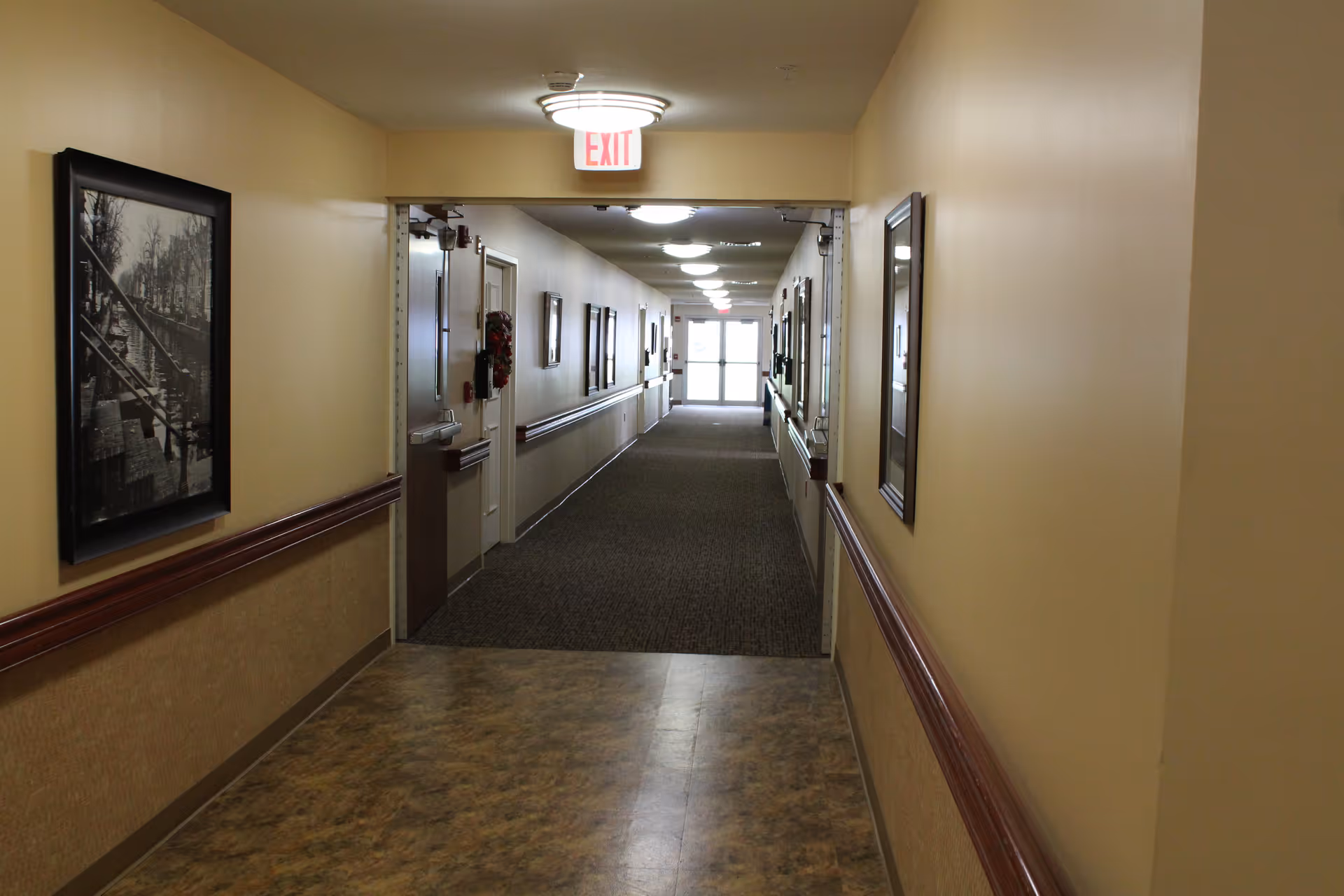 A long, well-lit indoor hallway in a senior living facility with beige walls, framed black and white photographs on the walls, and handrails on both sides. The floor transitions from a patterned tile to carpet as the hallway extends towards a glass exit door at the far end.