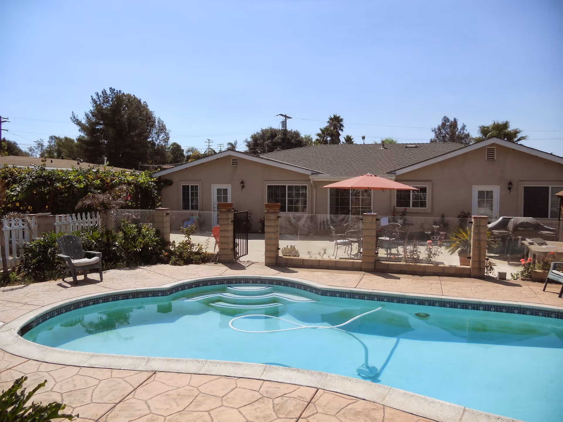 Backyard swimming pool and patio area with lounge chairs, an umbrella, and a single-story building in the background.